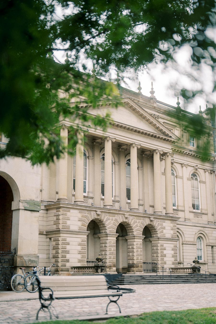 Historic building with classical architecture featuring columns and arches, surrounded by greenery and a cobblestone path with a bench and bicycles in the foreground.