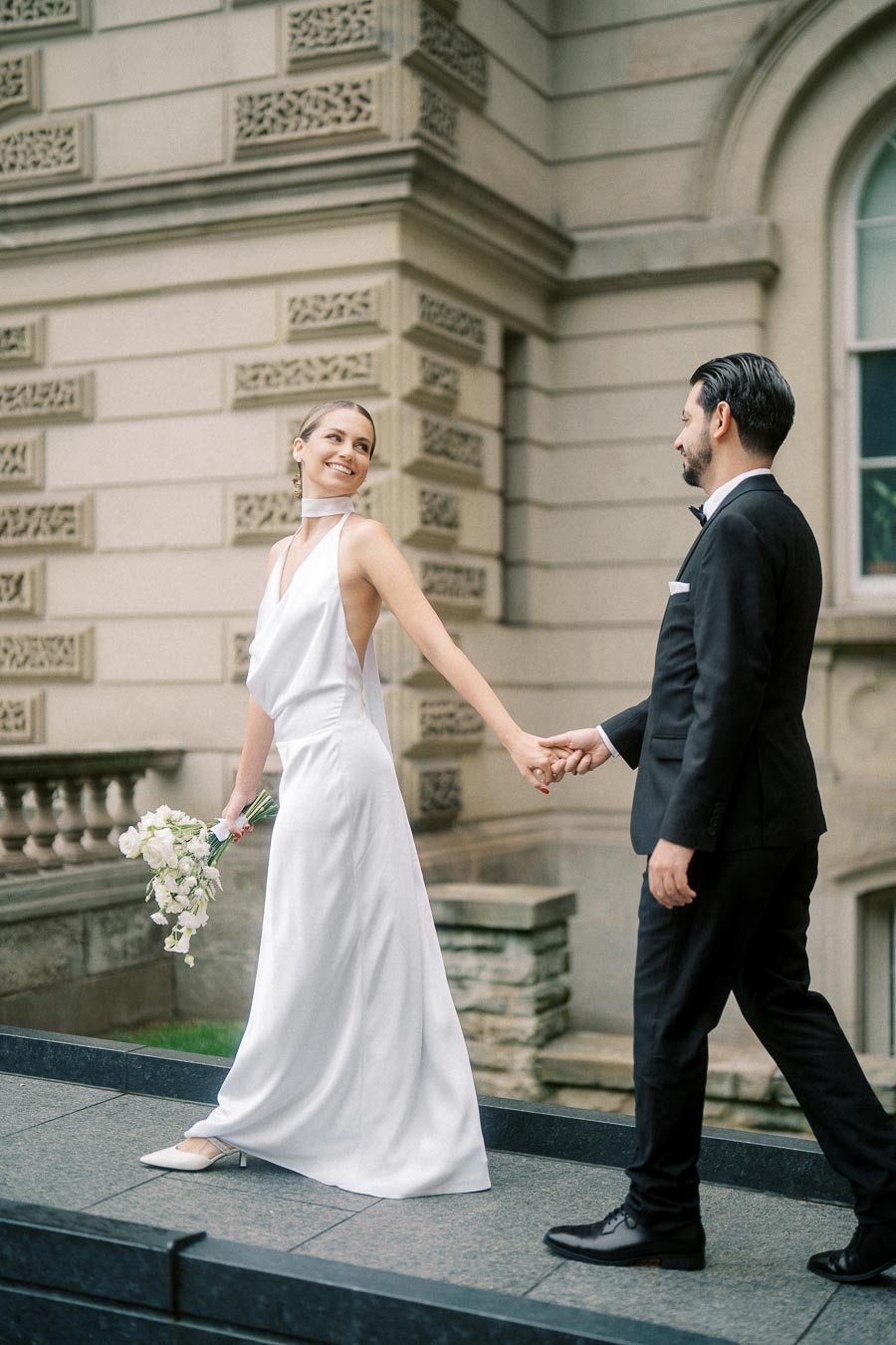 Bride in elegant white gown holding a bouquet, smiling and holding hands with groom in a black suit, walking in front of a historic architectural building.