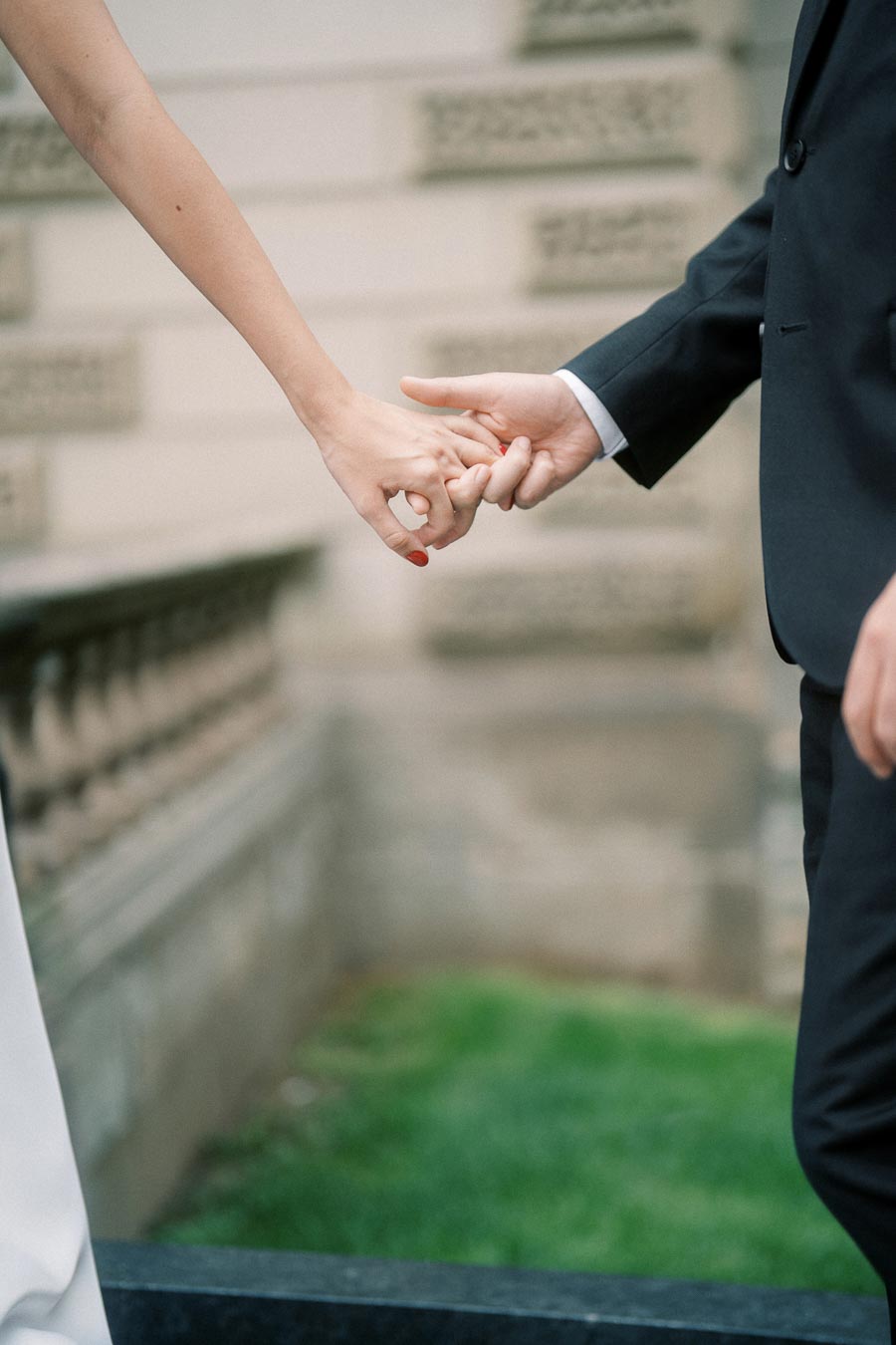 Close-up of a couple holding hands, with one wearing a black suit and the other a white outfit, symbolizing love and togetherness in a romantic outdoor setting.