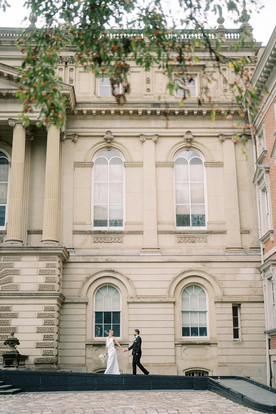 Elegant couple walking hand in hand outside a historic building with grand architectural details, including tall columns and arched windows.