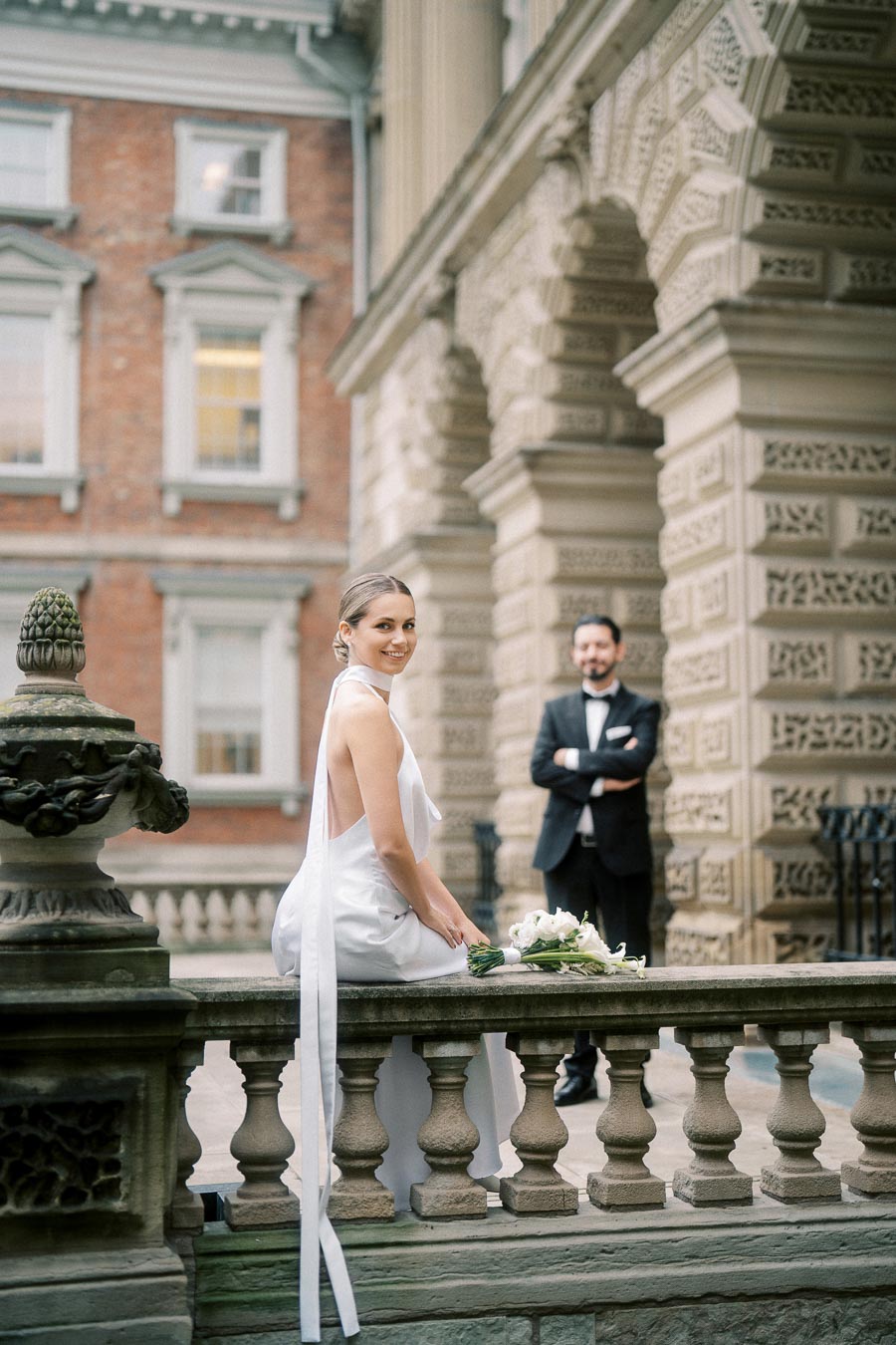 Bride in elegant white gown sitting on stone railing with bouquet, groom standing in the background wearing a tuxedo, with historical architecture in the backdrop.