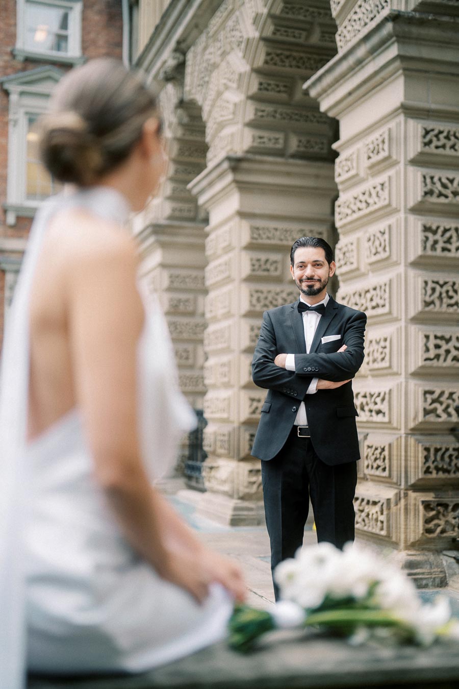 A groom in a black tuxedo and bowtie smiling with arms crossed, standing in front of an ornate stone building, while a bride in a white dress with her back to the camera, holding a bouquet of white flowers, sits in the foreground.