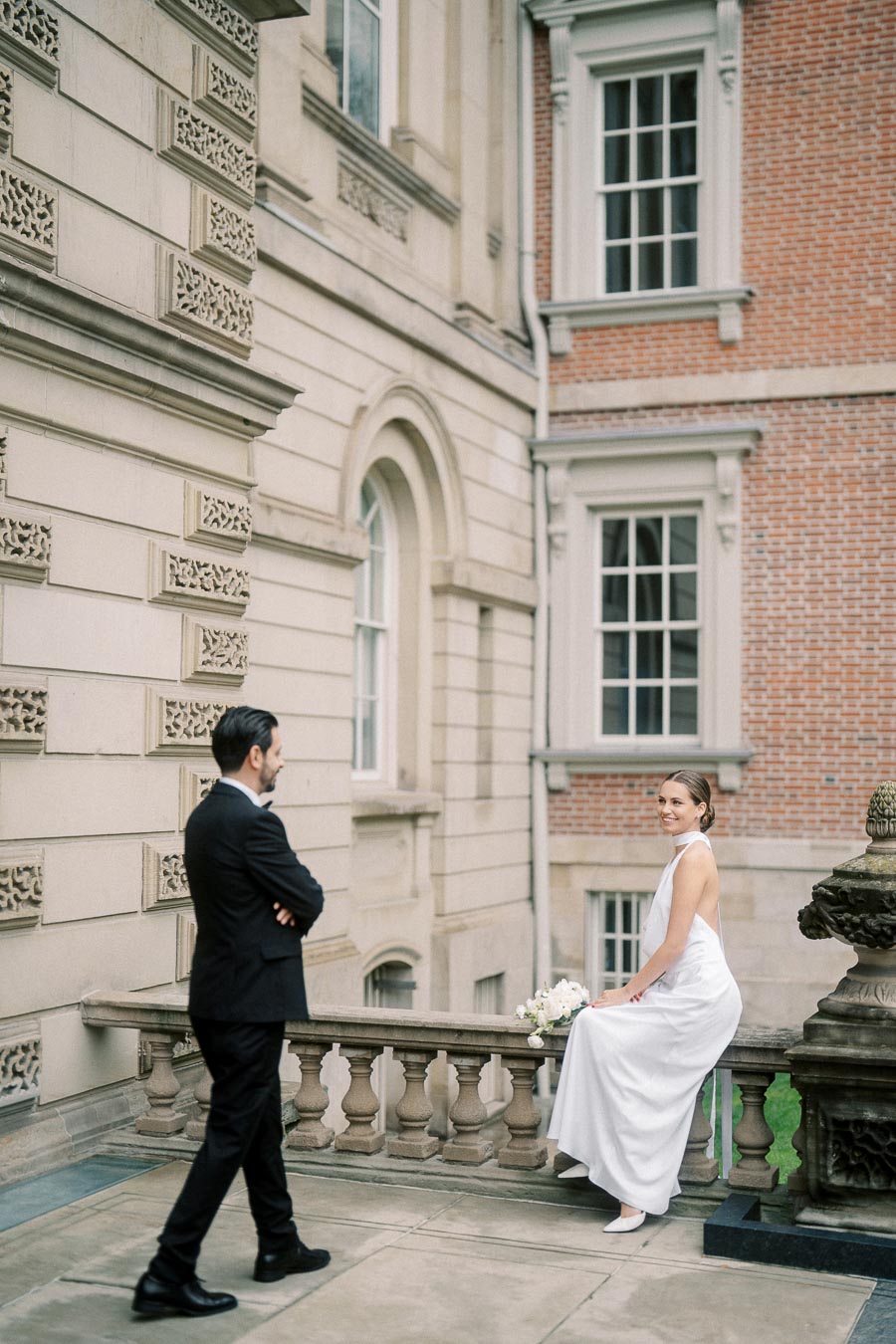A bride in a white wedding dress sits on a stone balustrade holding a bouquet, while a groom in a black suit stands nearby against a backdrop of an elegant, historic building with detailed architecture and large windows.