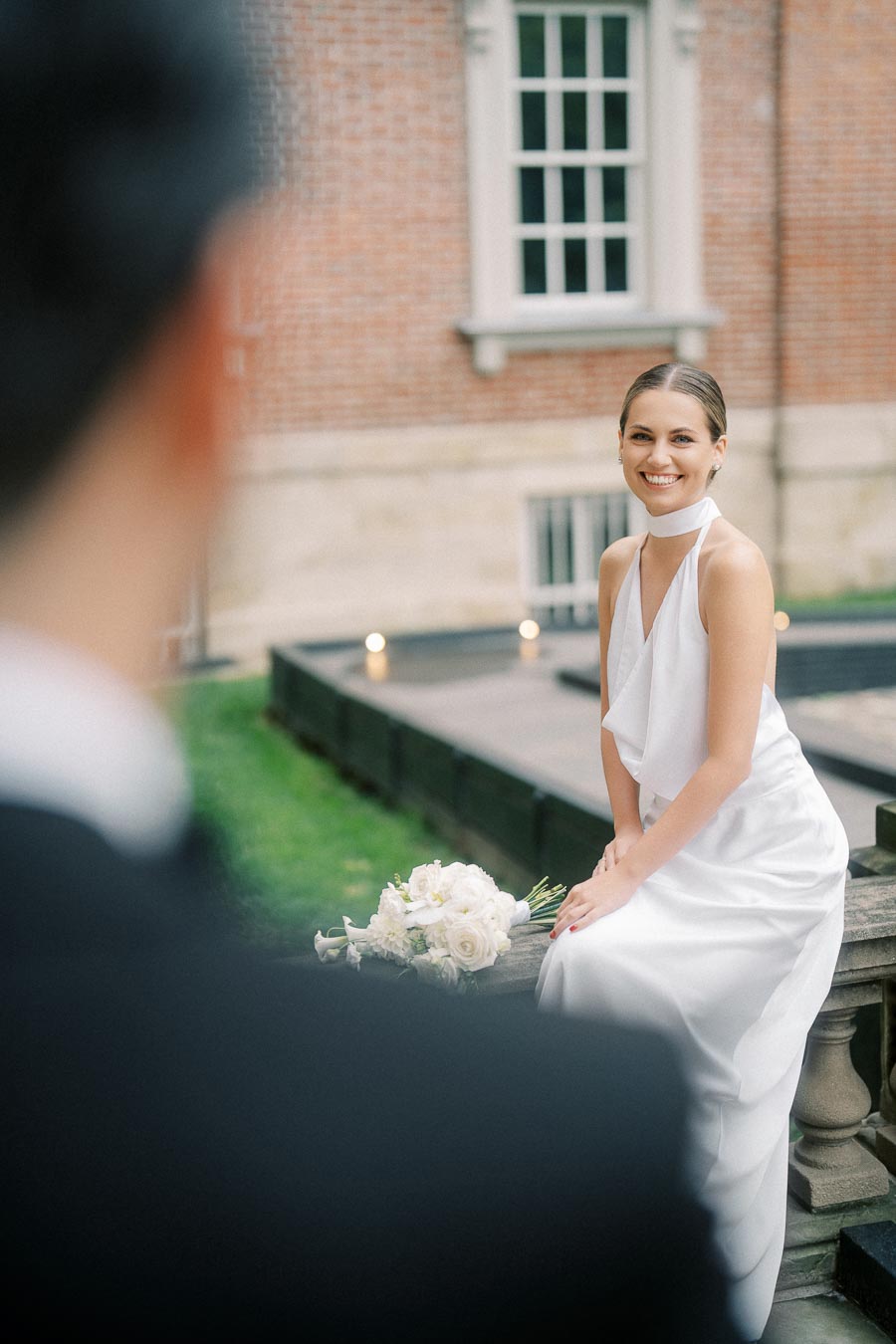 Bride in elegant white dress smiling during outdoor wedding photoshoot, holding bouquet of white roses, with historic brick building in background.