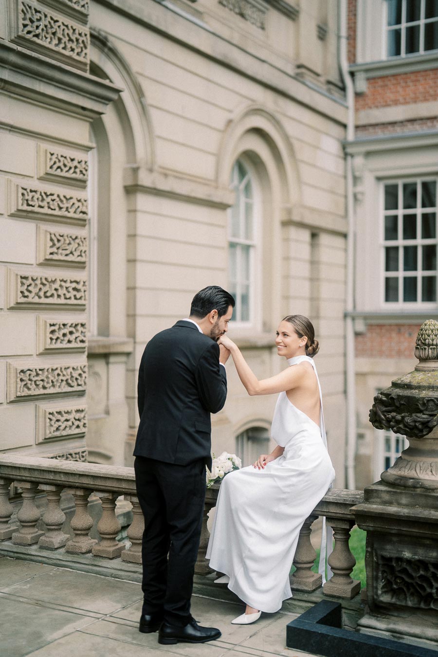 A bride in a white dress sits on a stone railing holding a bouquet, smiling as a groom in a black suit kisses her hand, with an ornate building in the background.