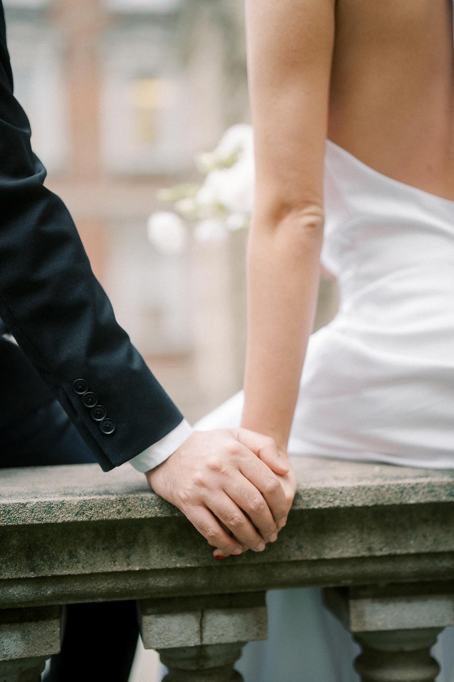 A bride and groom holding hands on a stone railing, showcasing wedding attire details with an elegant backdrop.
