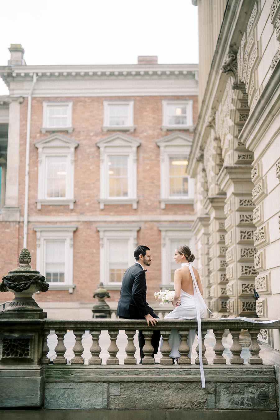 A bride and groom on a stone balcony wearing wedding attire, with the bride holding a bouquet of flowers. They are set against the backdrop of an elegant historic building, featuring detailed architecture and large windows.