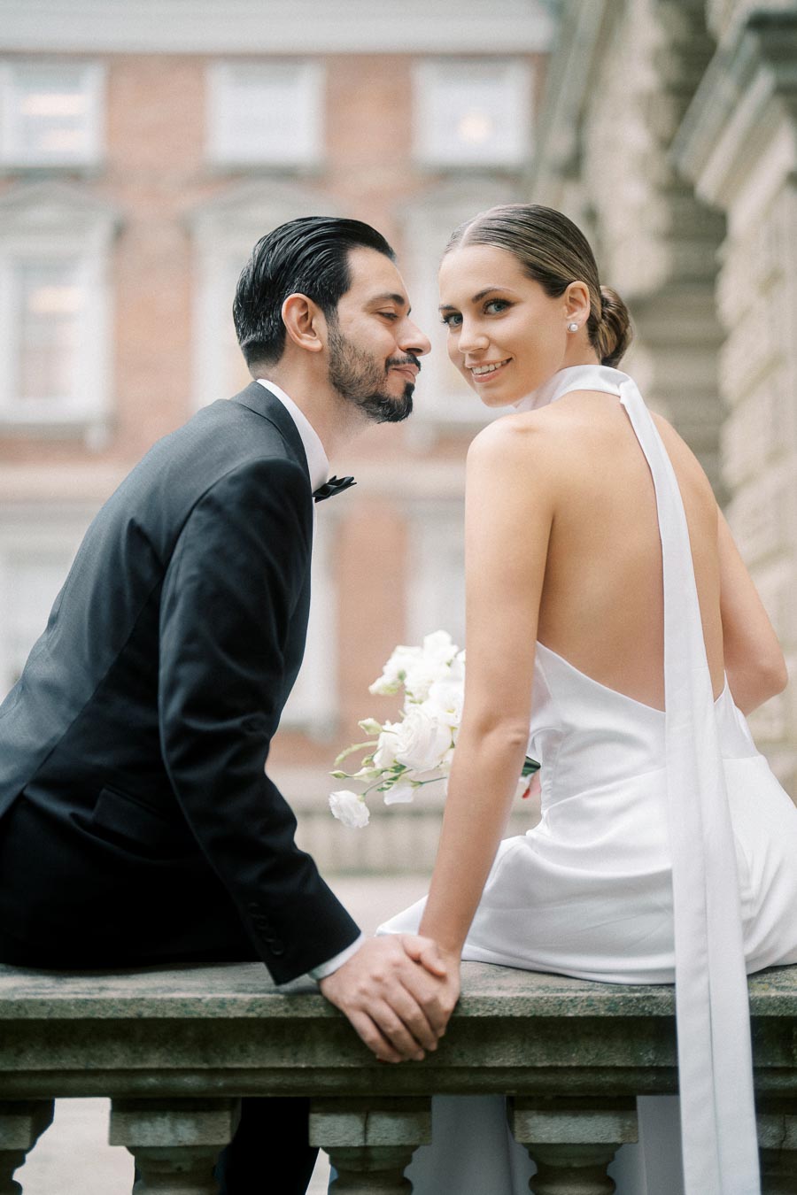 A couple dressed formally, with the woman in a white gown and the man in a black suit, sitting together on a stone ledge, holding hands and smiling. The background features a building with intricate architecture, suggesting a romantic occasion such as a wedding or engagement.
