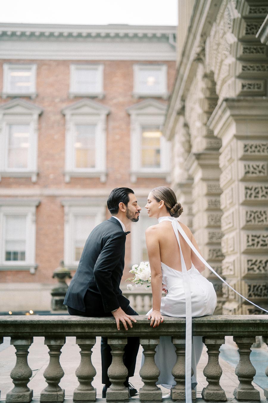 Elegant couple in formal attire sharing a romantic moment on a historic building balcony, with architectural details and a backdrop of large windows.