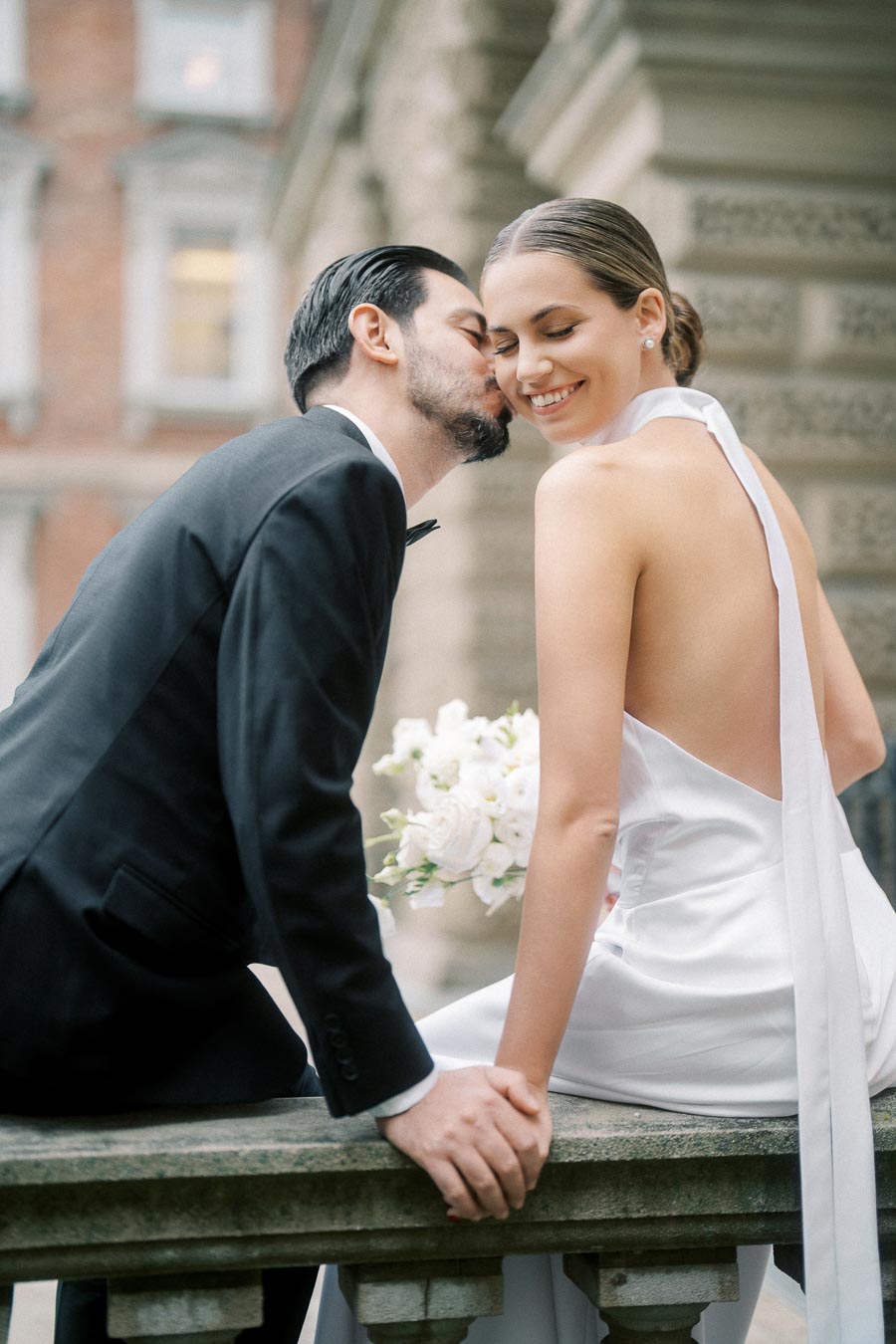 A bride in a white halter wedding dress smiles while sitting on a stone ledge, holding a bouquet of white flowers. A groom in a black tuxedo whispers affectionately in her ear, with an elegant architectural background.