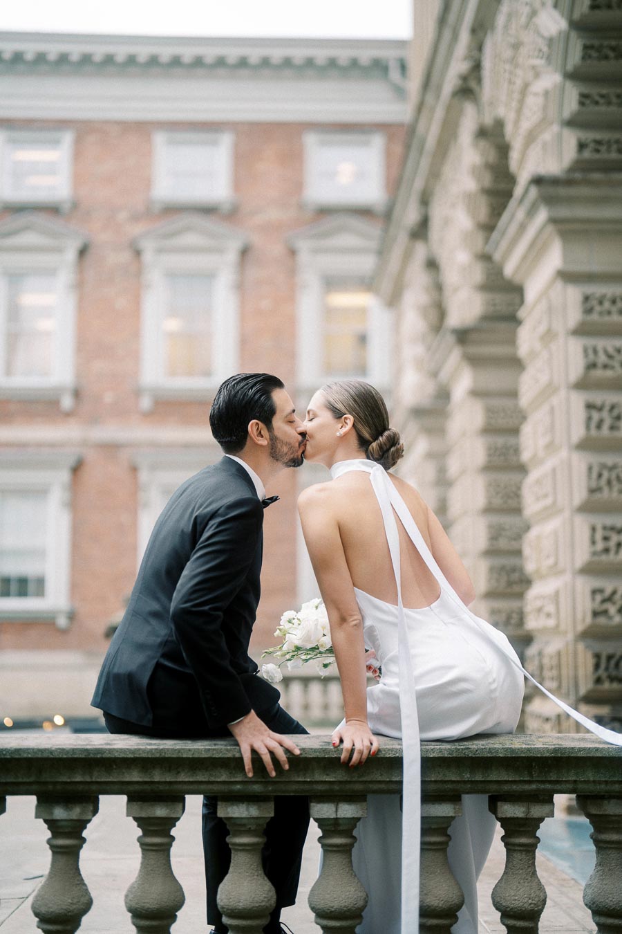 Bride and groom sharing a kiss in elegant wedding attire while sitting on an outdoor balustrade, surrounded by historic architecture.