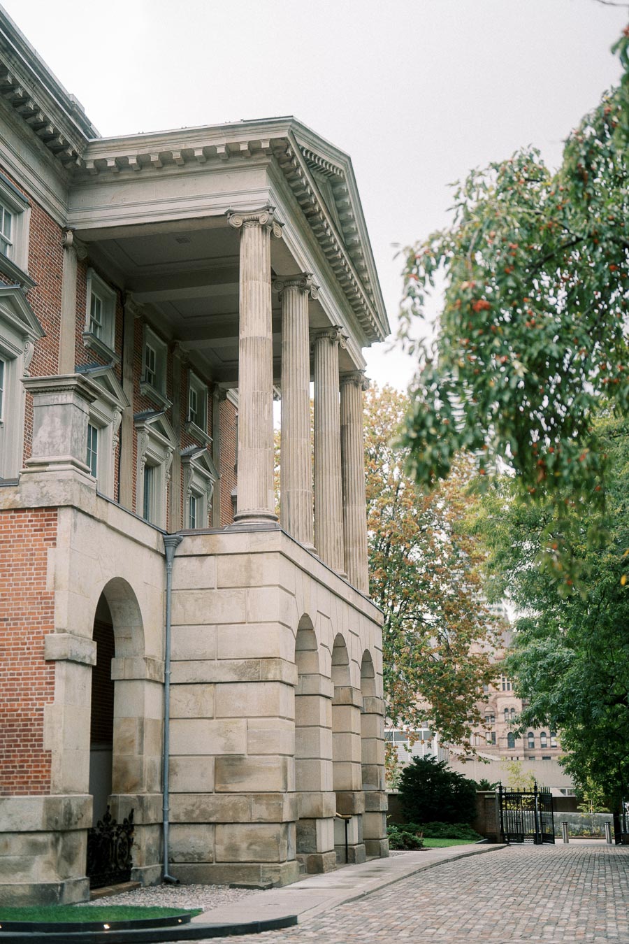 Classic architecture of a historic building with stone columns and arches amidst a tree-lined walkway and cobblestone path, showcasing a blend of traditional design and natural surroundings.
