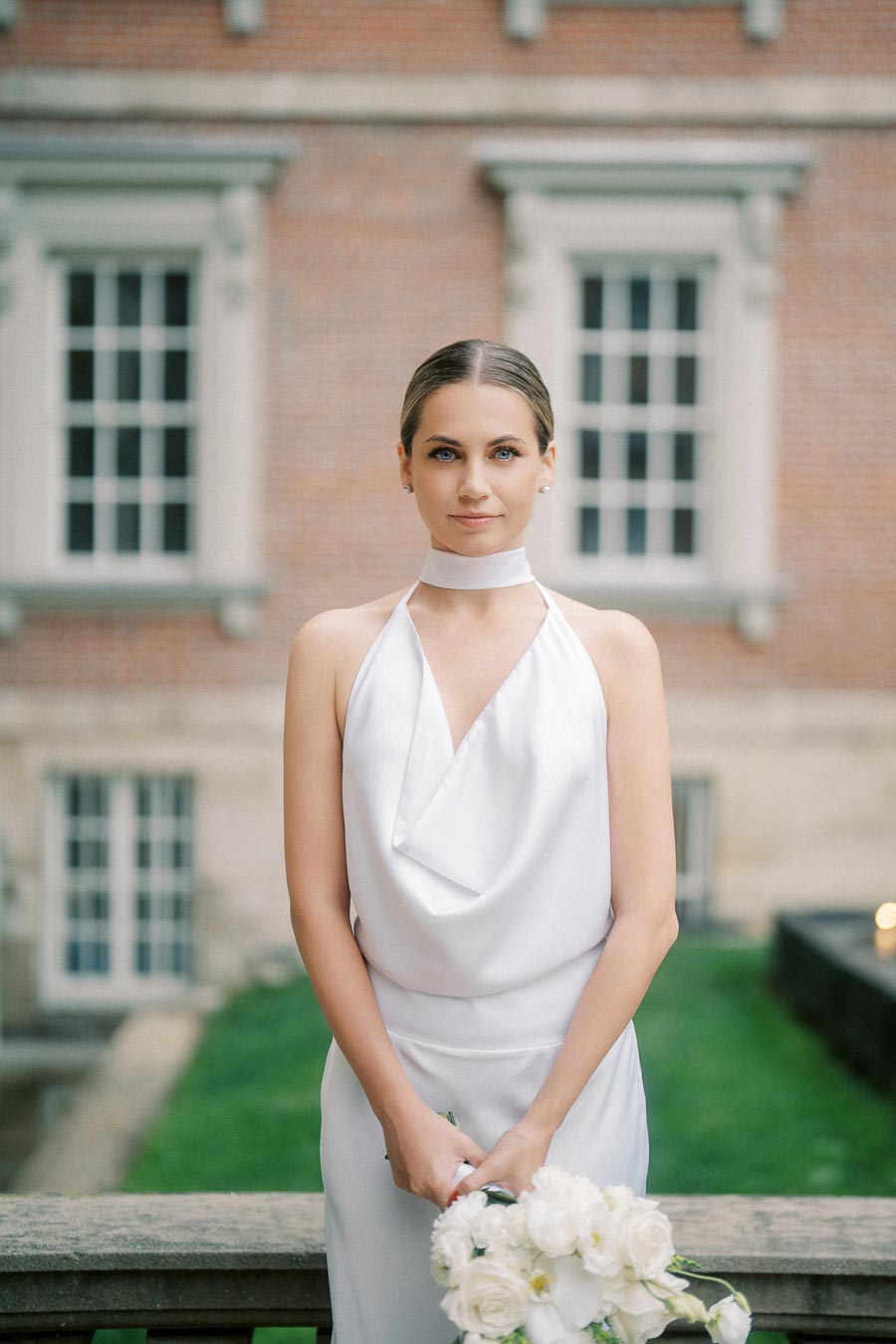 A bride in an elegant white halter-neck wedding dress holding a bouquet of white roses, standing in front of a historic brick building with large windows.
