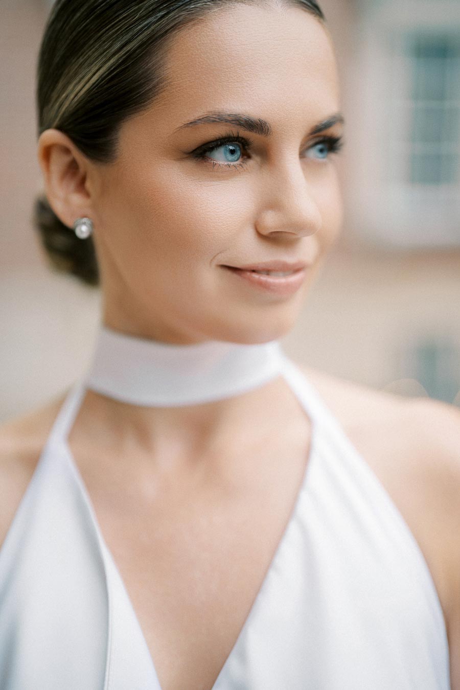 Elegant woman in bridal attire with a white halter dress and pearl earrings, showcasing a serene smile and sophisticated hairstyle, outdoors.