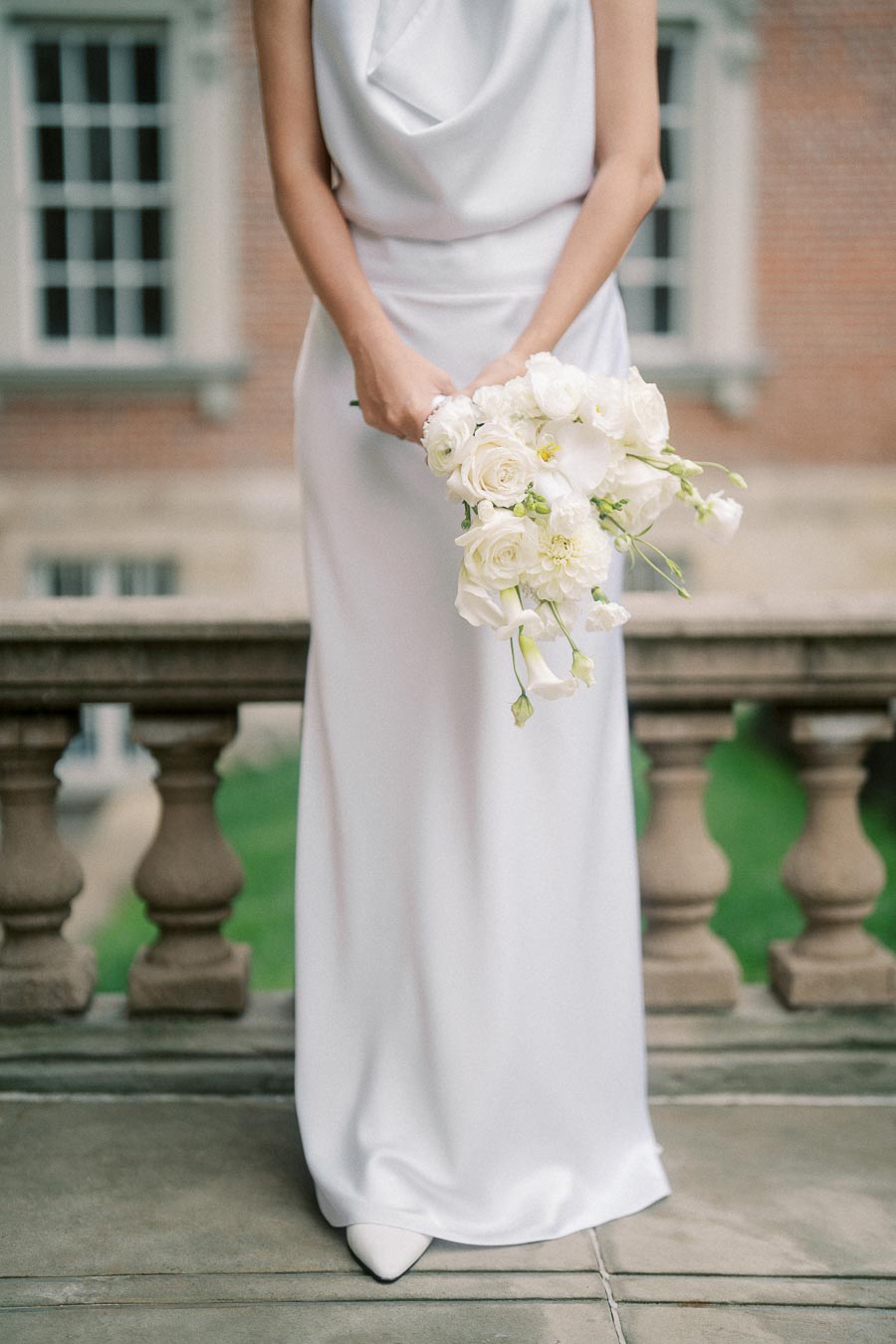 Elegant bride in a simple white wedding dress holding a bouquet of white flowers, standing by a stone balustrade with a historic building backdrop.