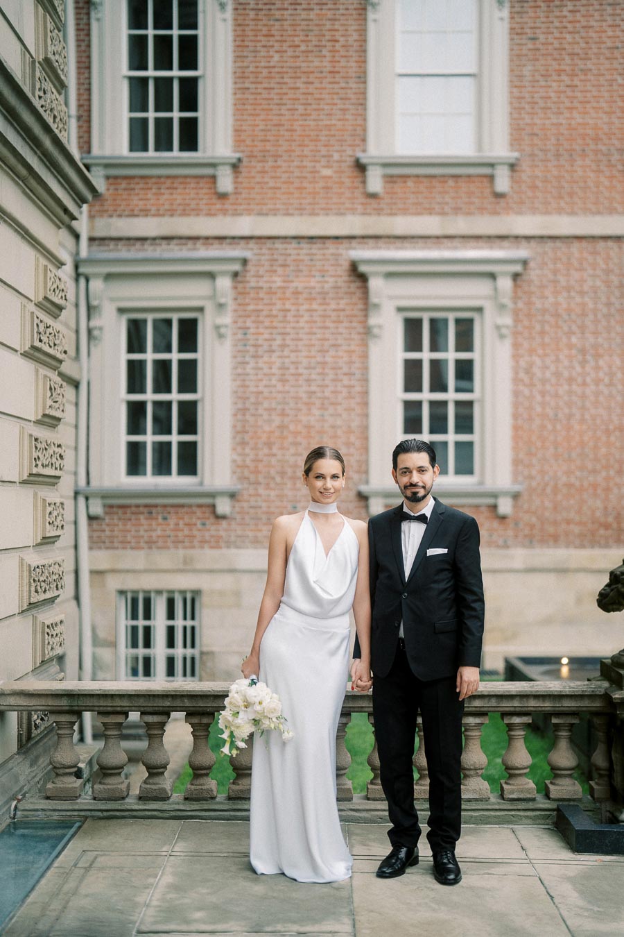 Elegant couple posing together on a stone balcony in front of a brick building, the woman in a white gown holding a bouquet and the man in a classic black tuxedo.