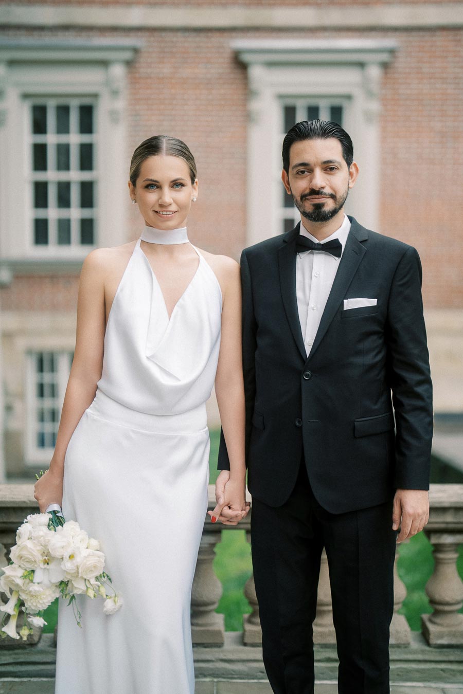 Elegant bride in a white gown holding a bouquet, standing next to a groom in a black tuxedo, outside a historic building.