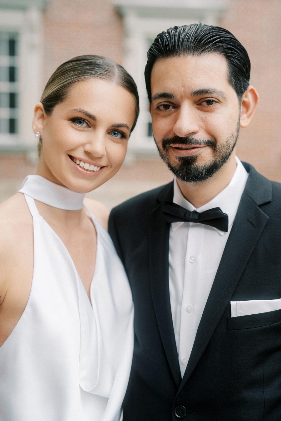 Elegant couple smiling at the camera during an outdoor wedding, with the woman in a white halter dress and the man in a black tuxedo with a bow tie.