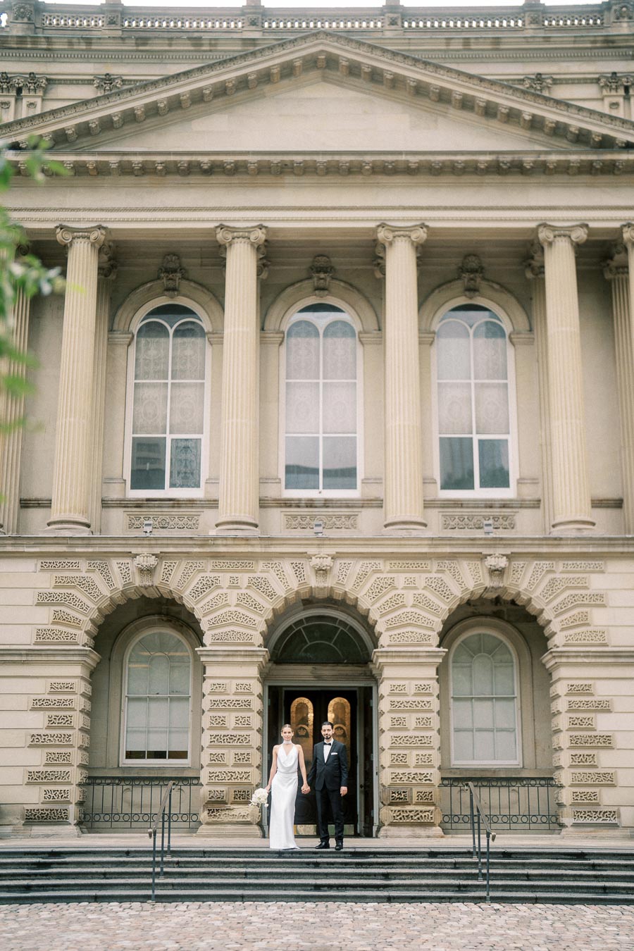 A bride and groom stand hand in hand on the steps of a historic building with large columns, elegant arches, and intricate architectural details. The bride is in a white dress, and the groom is in a black suit, both looking forward and smiling, capturing a timeless wedding moment.