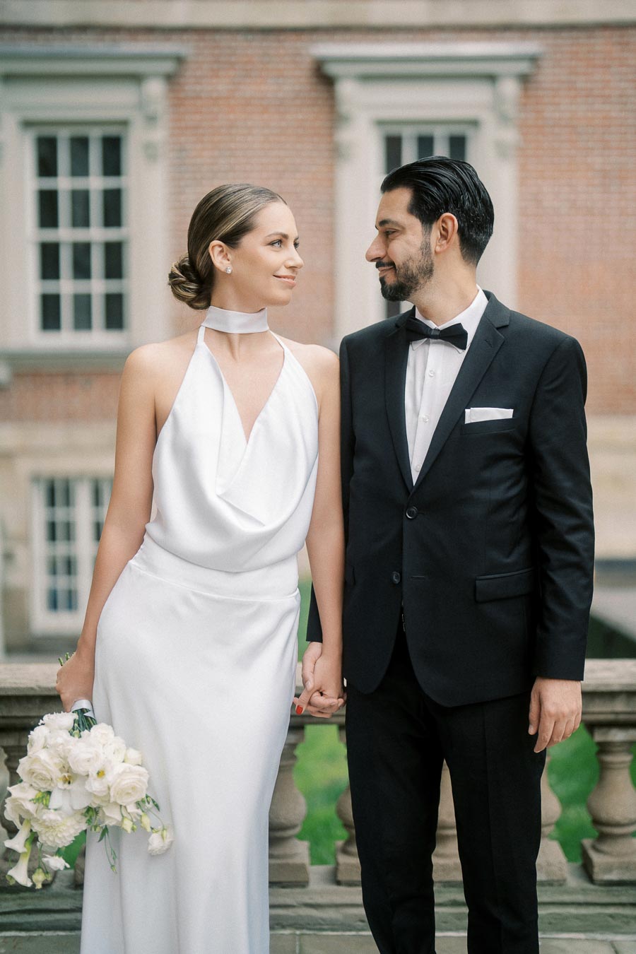 Elegant couple holding hands in formal attire, with the woman in a white wedding dress holding a bouquet of white flowers and the man in a black suit and bow tie, standing in front of a historic brick building.