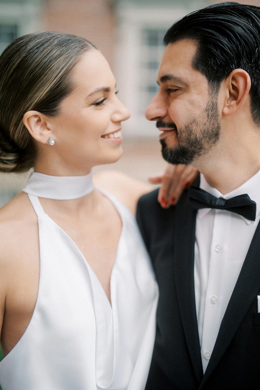 A bride in a white dress and groom in a black tuxedo smiling at each other during their wedding photoshoot.