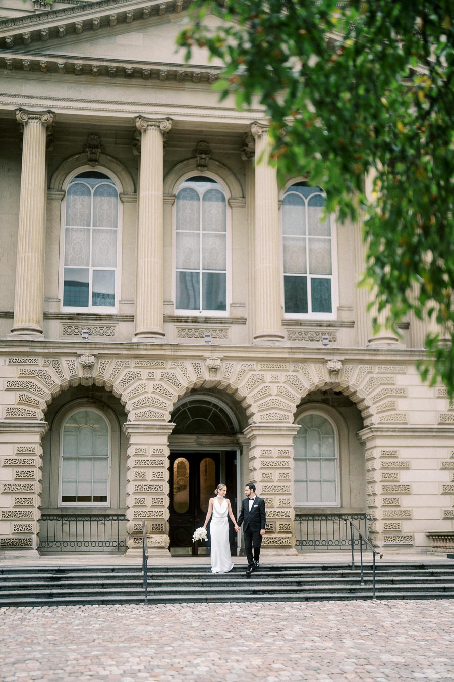 Elegant couple in wedding attire walking down the steps of a historic building with classic architectural columns.
