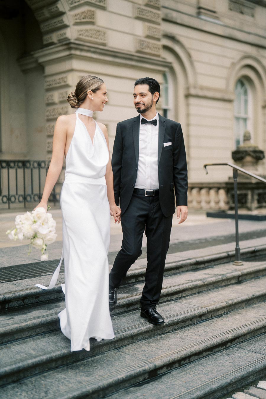 Elegant bride and groom walking down stone steps, with the bride holding a bouquet of white flowers, both smiling at each other, in front of an ornate building.