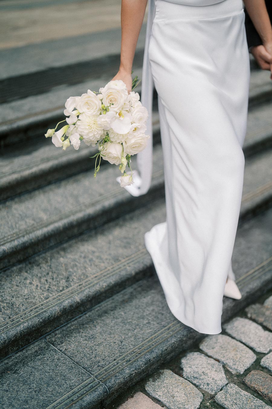 Bride holding a white floral bouquet while walking down stone stairs in an elegant wedding dress.