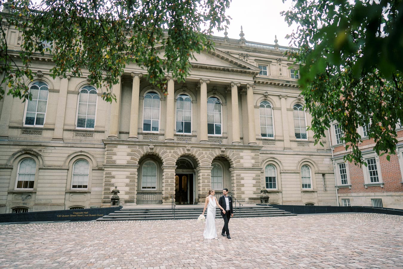 Wedding couple walking in front of a grand historic building with detailed stone architecture, columns, and arched windows, surrounded by cobblestone pavement and green foliage.