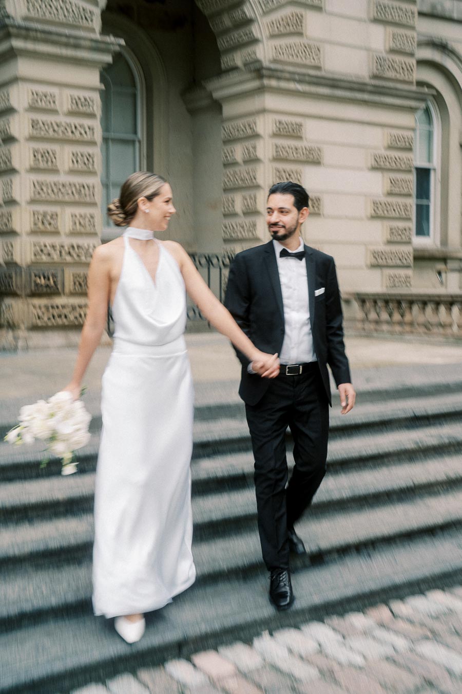 Elegant wedding couple walking down steps, the bride in a white gown holding a bouquet and the groom in a black suit, in front of a historic stone building.