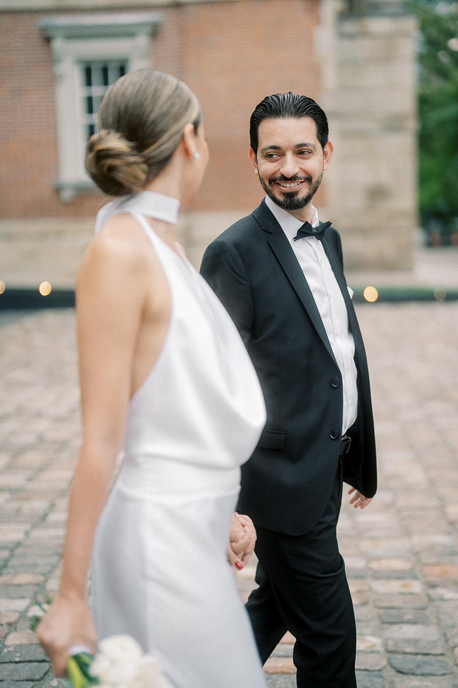 A bride in a white dress and a groom in a black suit share a joyful moment while holding hands outside on a cobblestone walkway.