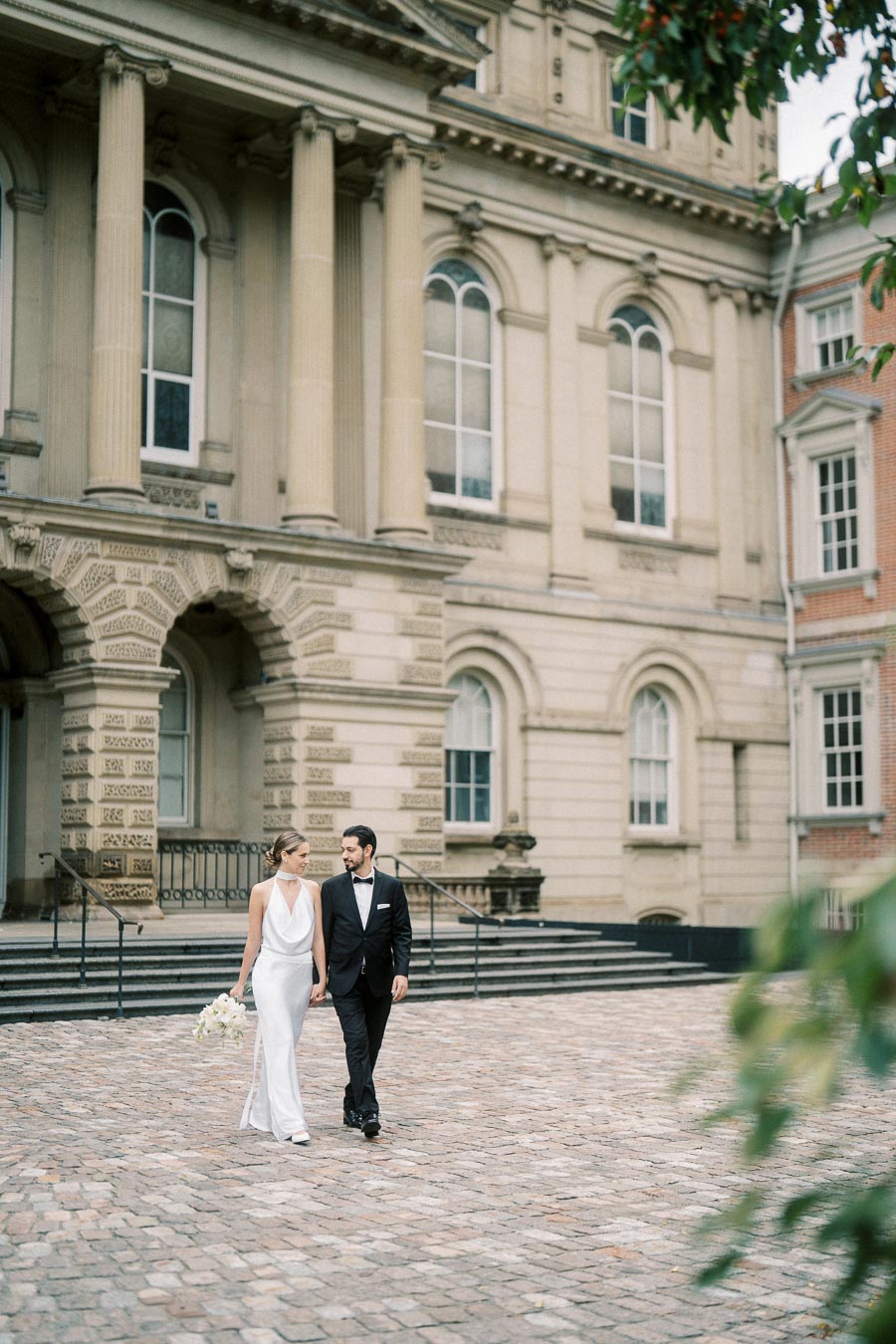 A bride in a white dress and a groom in a black tuxedo walking hand in hand in front of a historic building with tall columns and arched windows; the scene captures a romantic wedding moment.