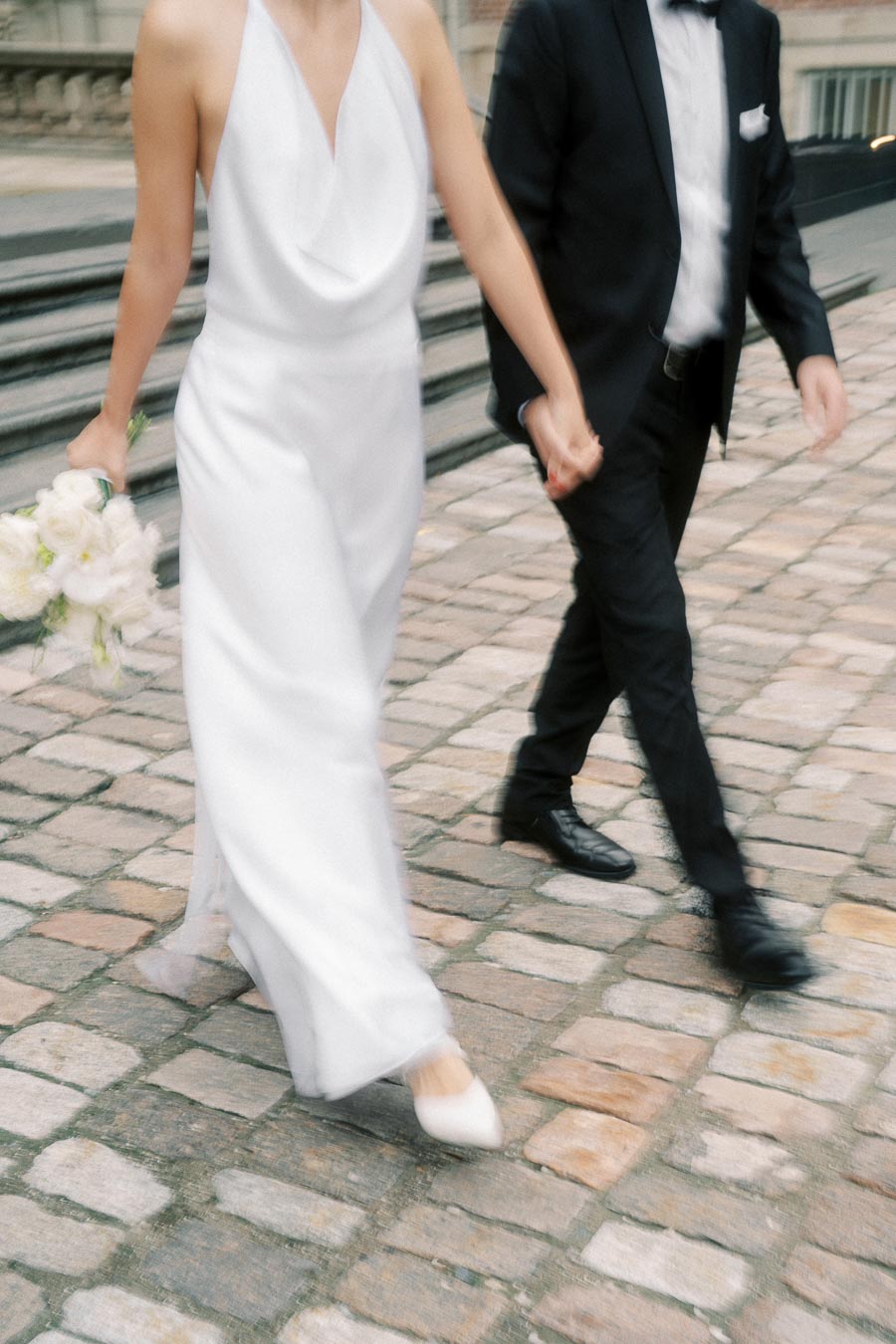 A bride in a white dress holding a bouquet walks hand in hand with a groom in a black suit on cobblestone pavement.