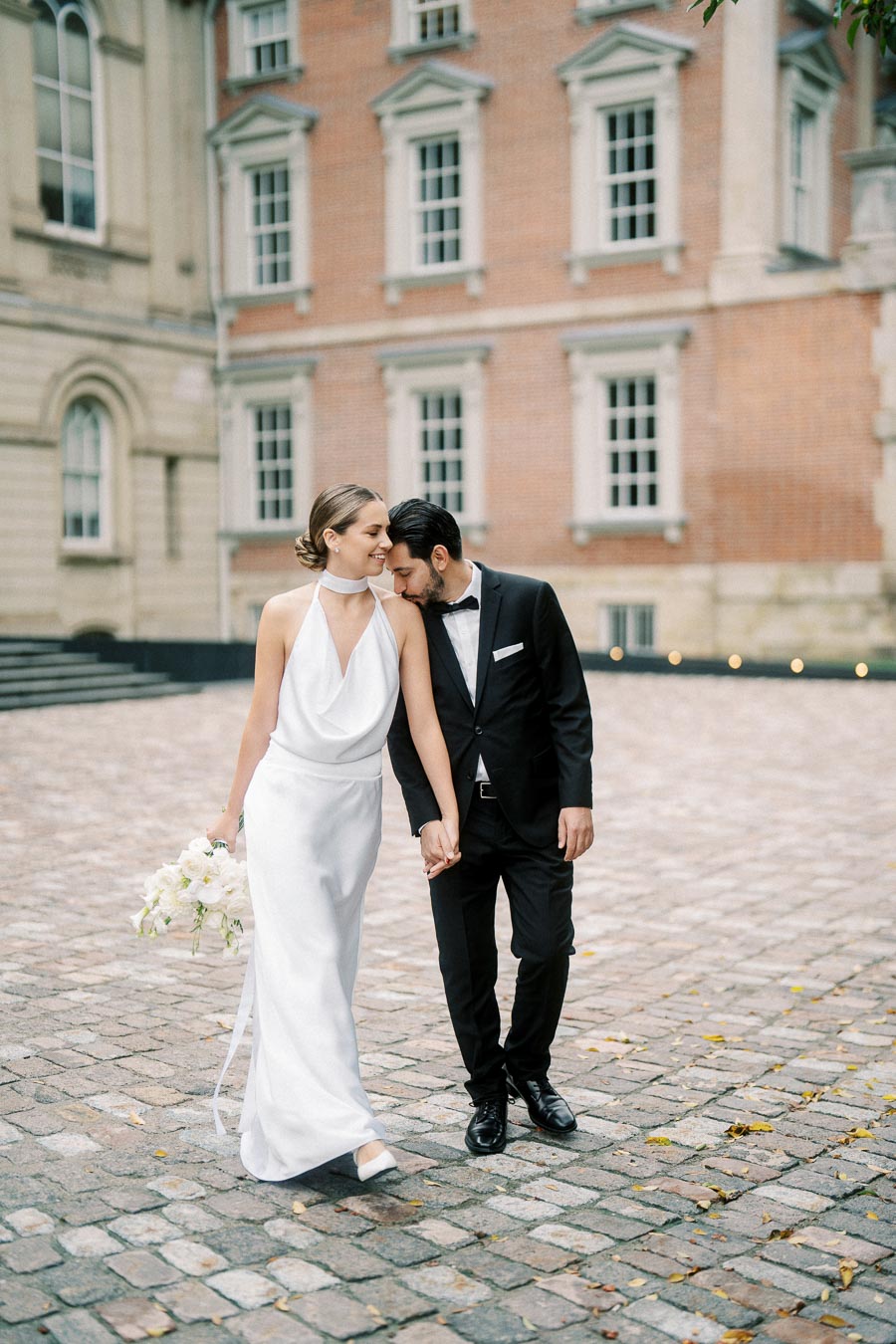 A bride in an elegant white gown and a groom in a classic black suit share a tender moment while walking hand in hand on cobblestone pavement, with a historic brick building in the background.