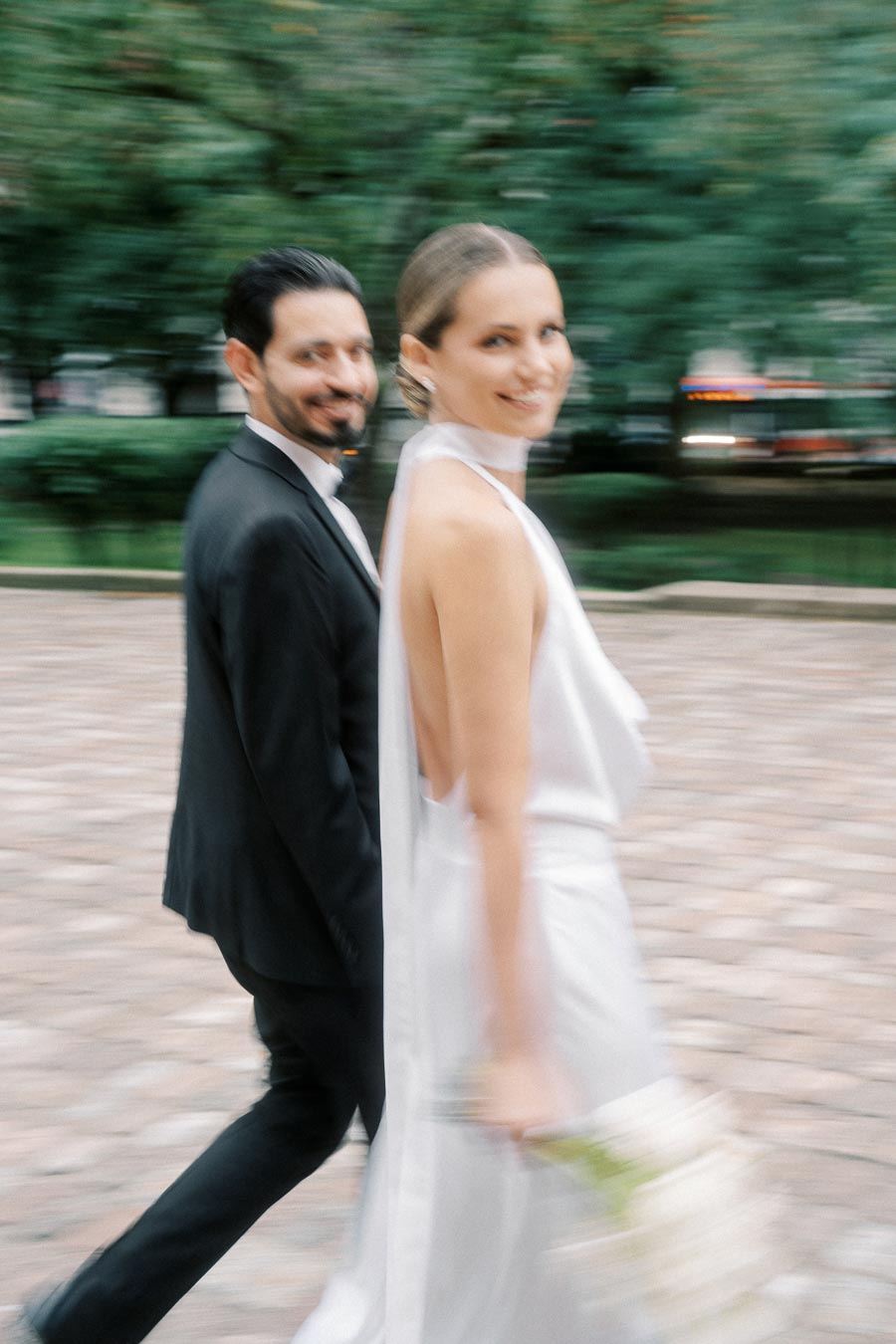 A couple in elegant attire walking outdoors, with the woman in a white dress holding a bouquet, and the man in a dark suit. The background appears blurred, suggesting motion in a park setting.