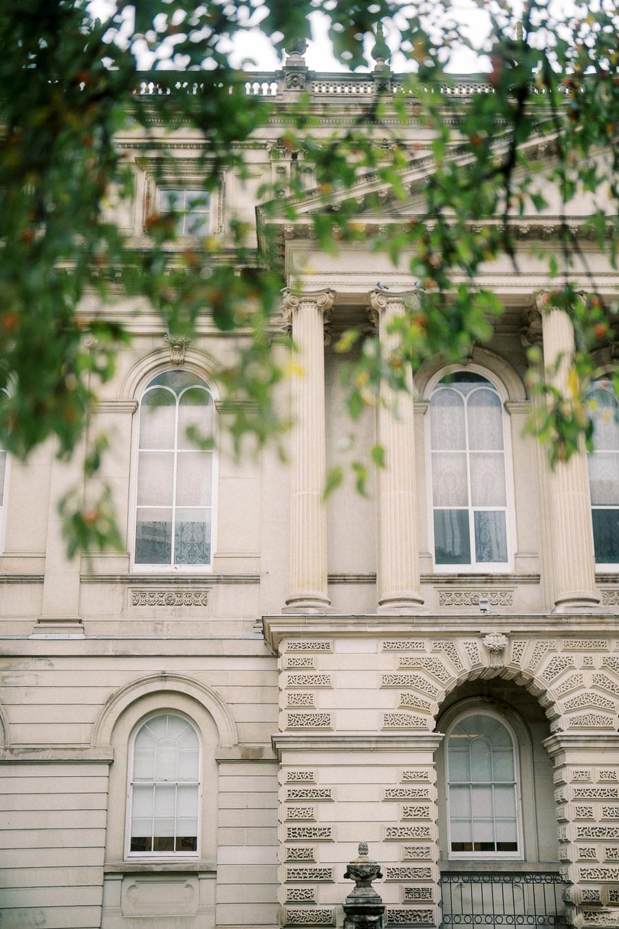 Elegant neoclassical building facade with tall columns and arched windows, partially obscured by leafy branches in the foreground.
