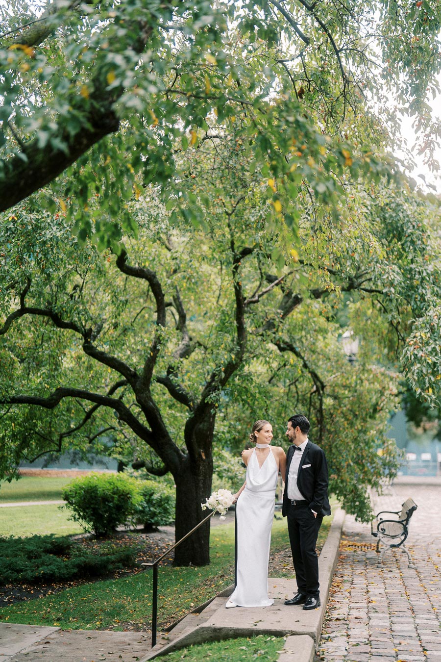 Elegant bride and groom standing together in a lush green park under a large tree, the bride holding a bouquet of flowers, both wearing formal wedding attire.