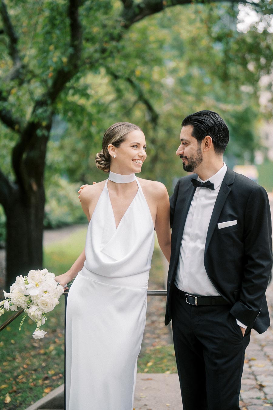 Elegant bride in a white gown and groom in a black tuxedo smiling at each other, holding a bouquet of white flowers, standing outdoors under lush green trees.