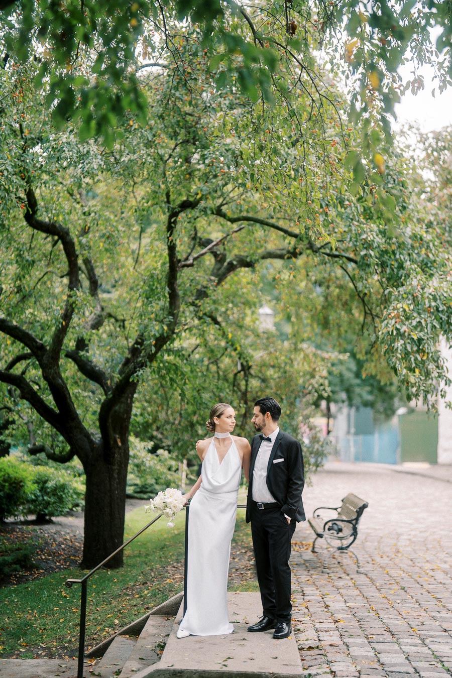 Elegant couple standing near a tree in a serene park, the bride in a white gown holding a bouquet and the groom in a black tuxedo, with cobblestone path and lush greenery in the background.