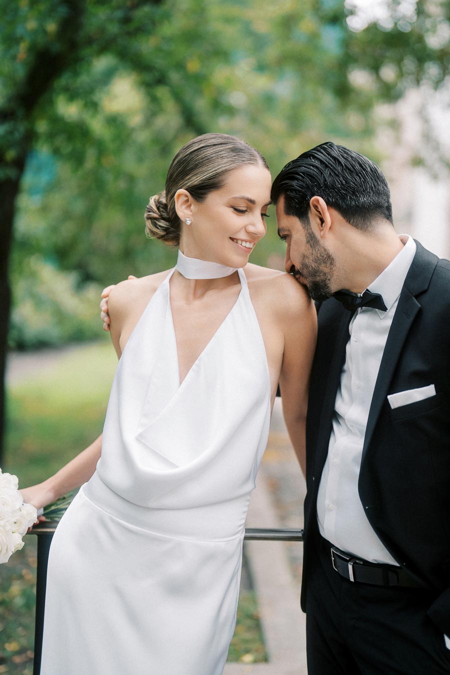 Elegant couple embracing in outdoor park setting, wearing a sleek white bridal gown and classic black tuxedo, surrounded by lush greenery.
