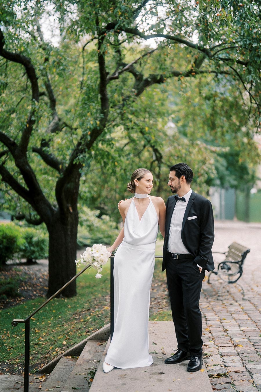 A bride in a white halter-neck gown and a groom in a black tuxedo smiling at each other on a cobblestone path in a lush green park.