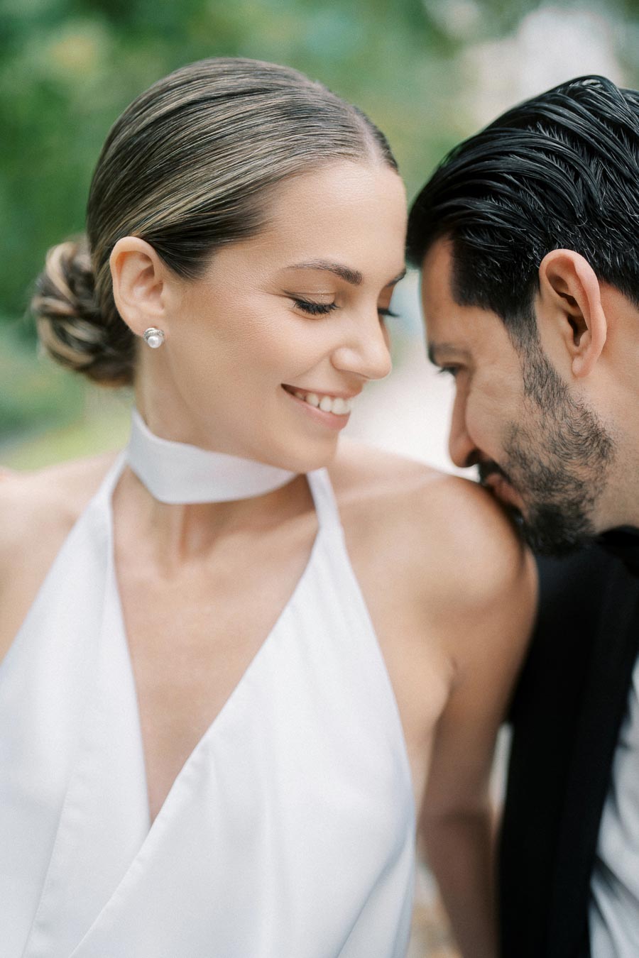 A happy couple smiling intimately, dressed in formal attire, surrounded by a blurred natural background.
