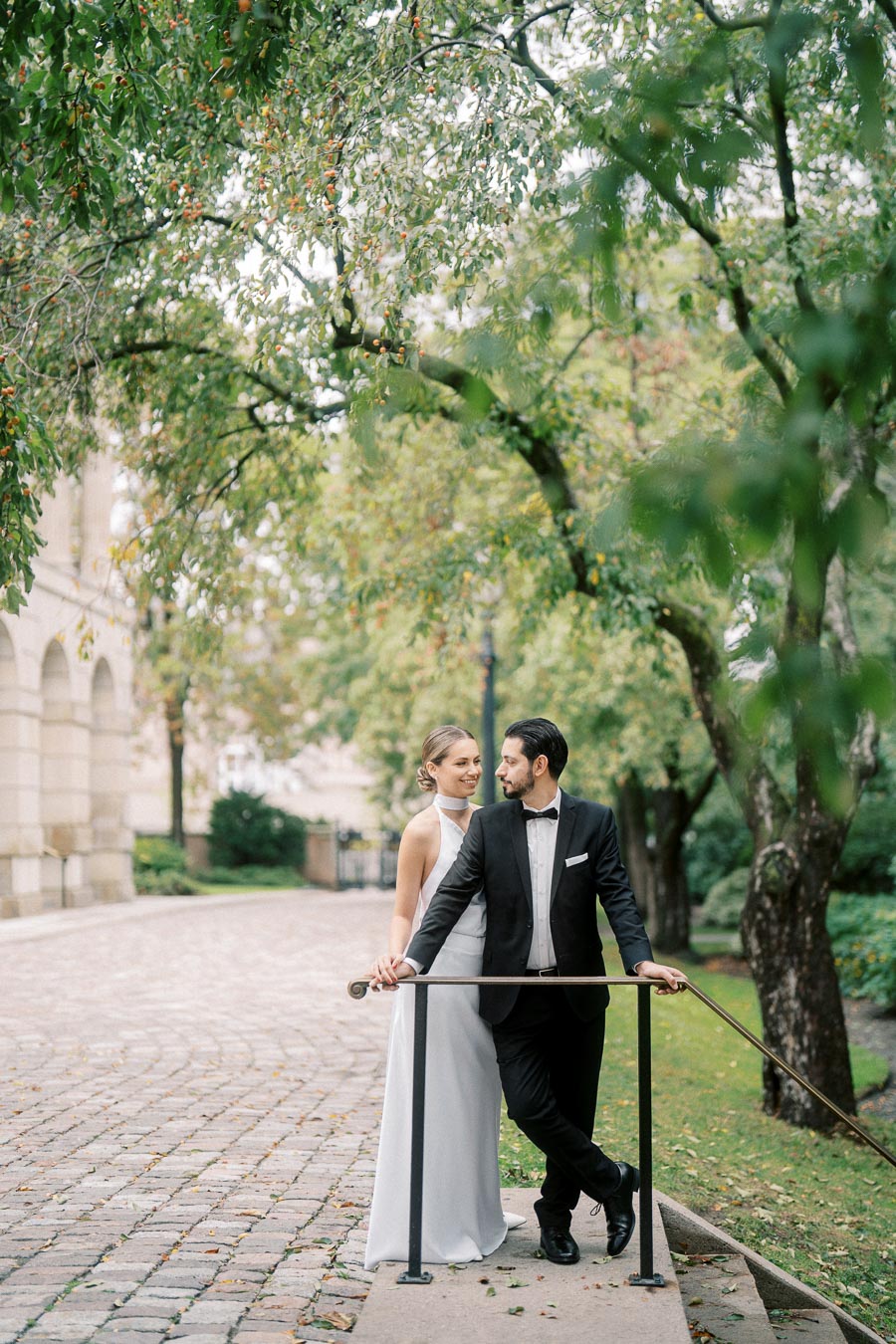 Elegant couple in formal attire standing by a pathway lined with lush trees, perfect for a wedding photoshoot backdrop.