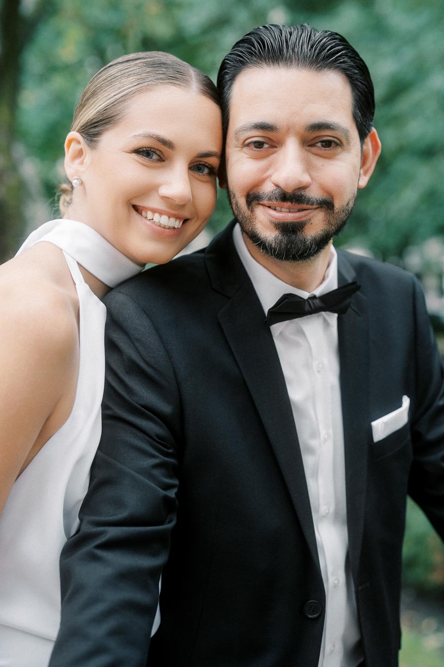 Smiling couple in elegant wedding attire posing outdoors with a natural green background.