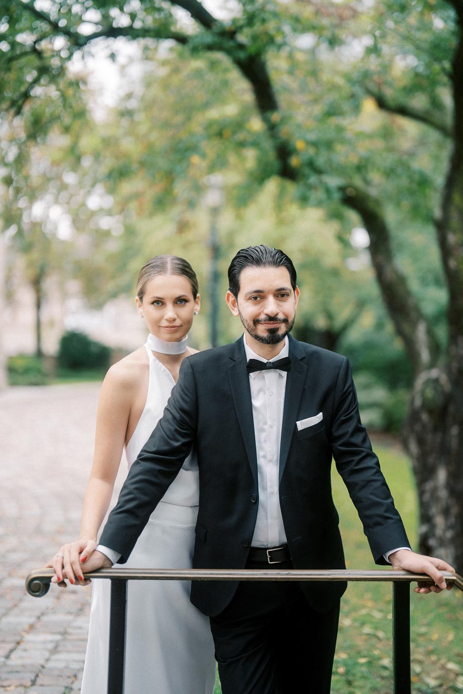 A bride in a white dress and a groom in a black tuxedo stand together in a scenic outdoor setting with lush greenery and a cobblestone path.