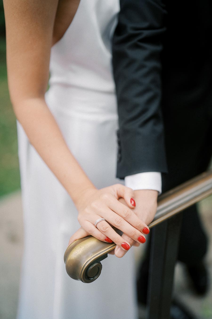 Bride and groom holding hands on a brass railing, showcasing wedding rings, with the bride wearing a white dress and red nail polish.
