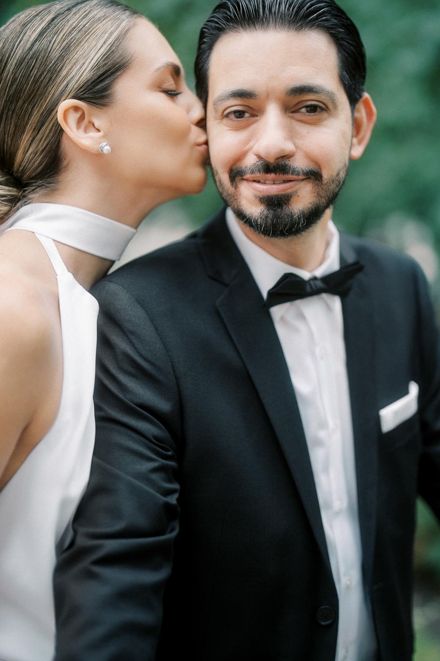 A woman in a white dress kisses a man in a black suit and bow tie, smiling outdoors with a blurred green background.