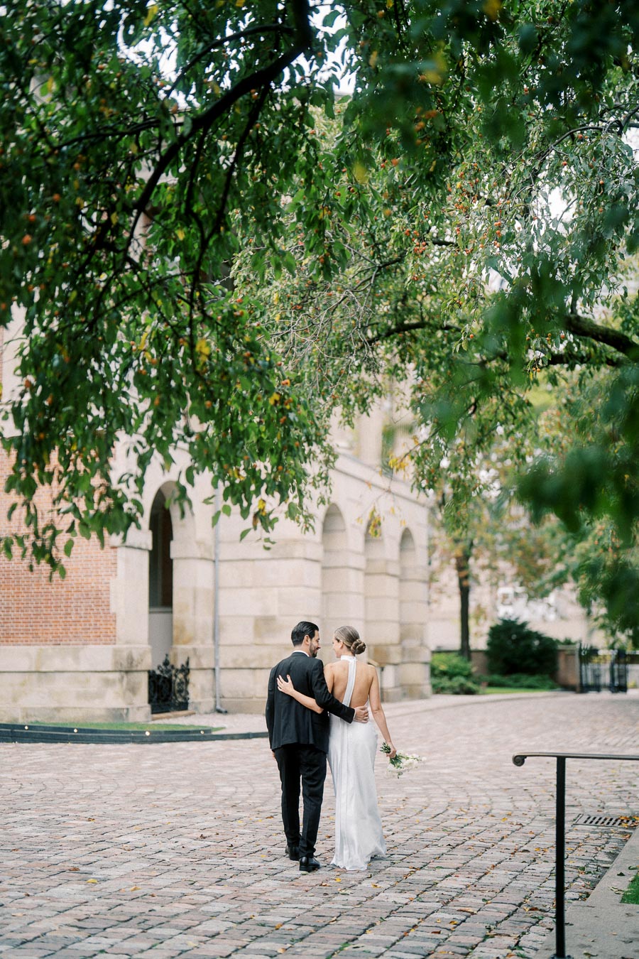 Wedded couple walking hand in hand on a cobblestone path under lush green trees beside a historic building, bride in white dress holding flowers, romantic outdoor scene.