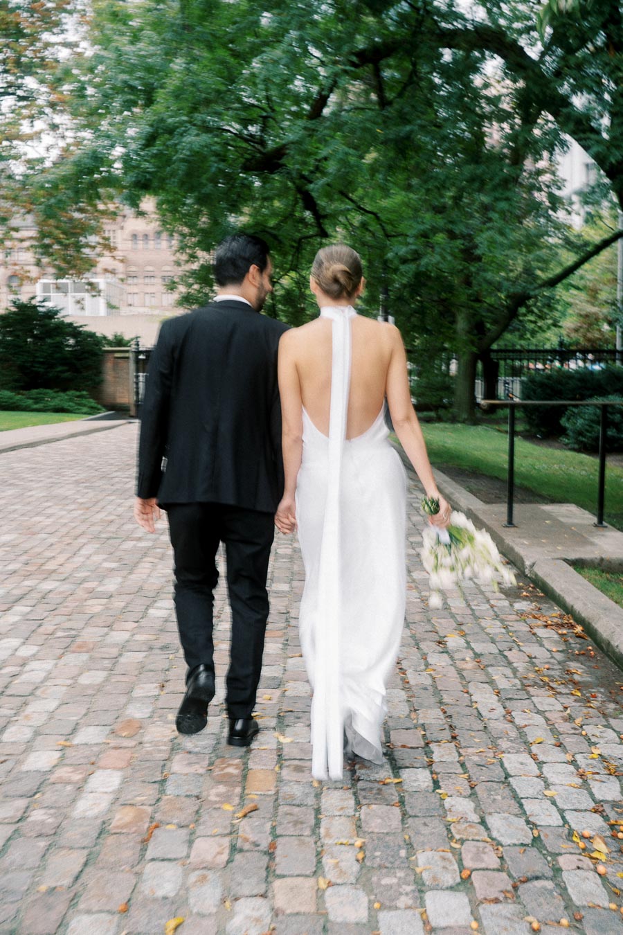 Couple walking hand in hand on a cobblestone path, the bride in a white backless gown holding flowers, with lush green trees and a historic building in the background, capturing a romantic wedding moment.