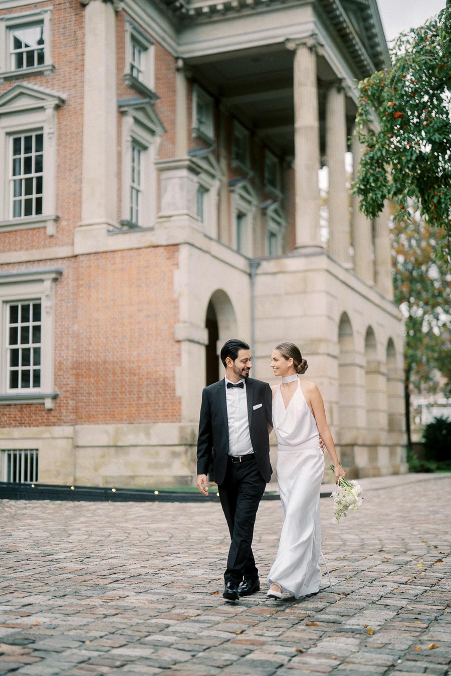 Elegant couple in formal attire walking hand in hand outside a historic brick building with columns, symbolizing romance and sophistication.